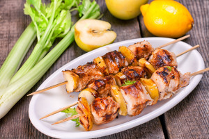 Three skewers of grilled chicken fillet in honey-mustard sauce with apples on a white plate on a wooden table